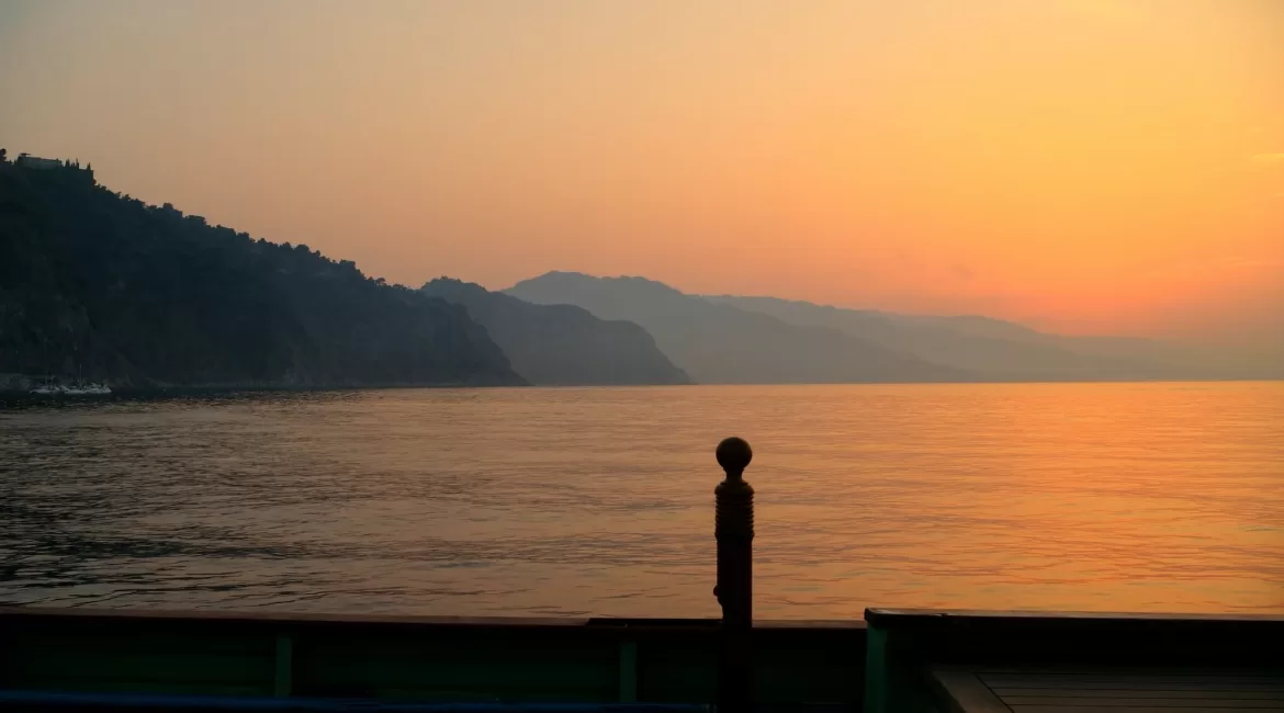 Golden sunset over the calm sea off Taormina seen from a boat deck, coastline in silhouette